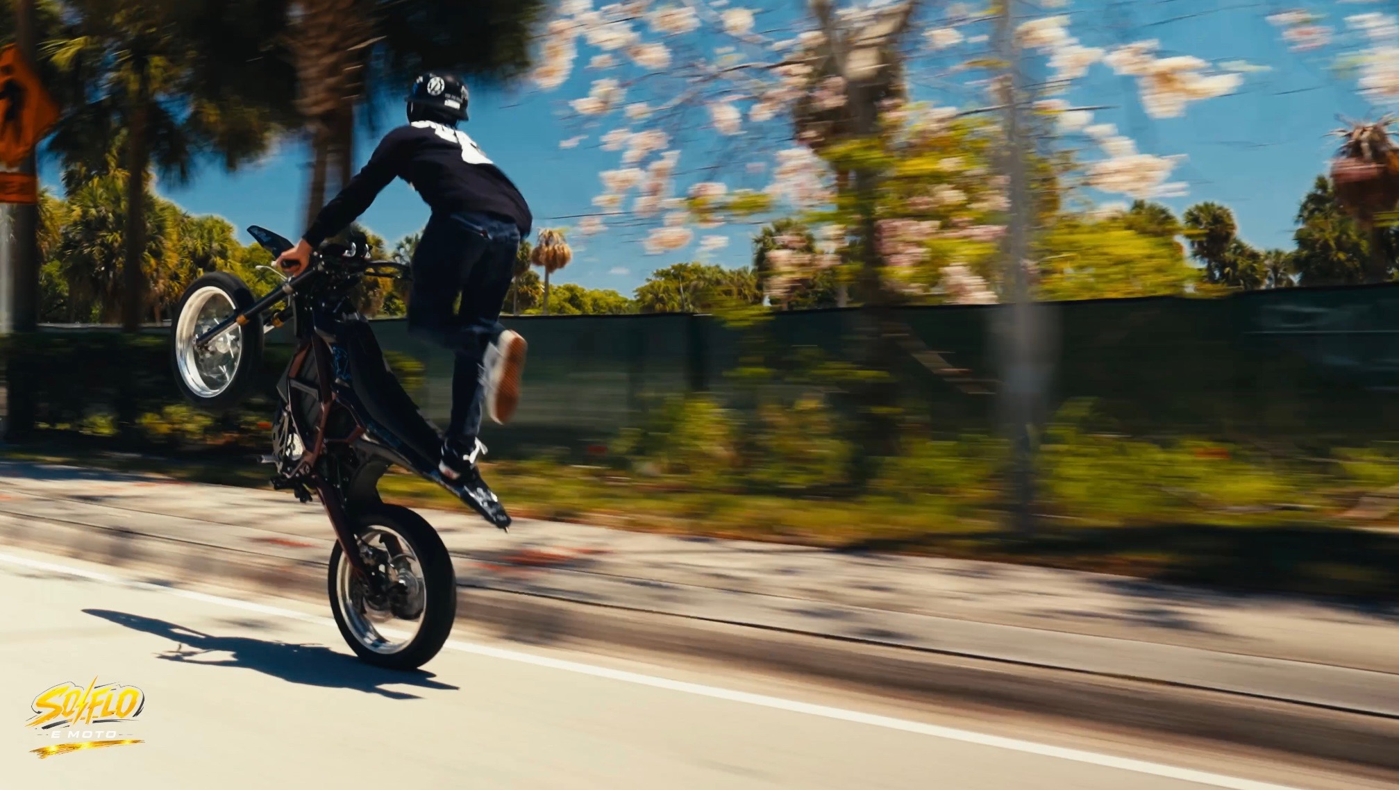 SoFlo E-Moto rider throwing a fast wheelie on a palm-lined street.
